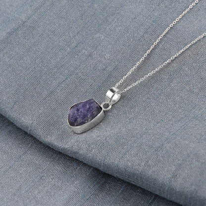 Raw blue tanzanite crystal pendant placed on fabric background showcasing natural rough cut and elegant bezel setting