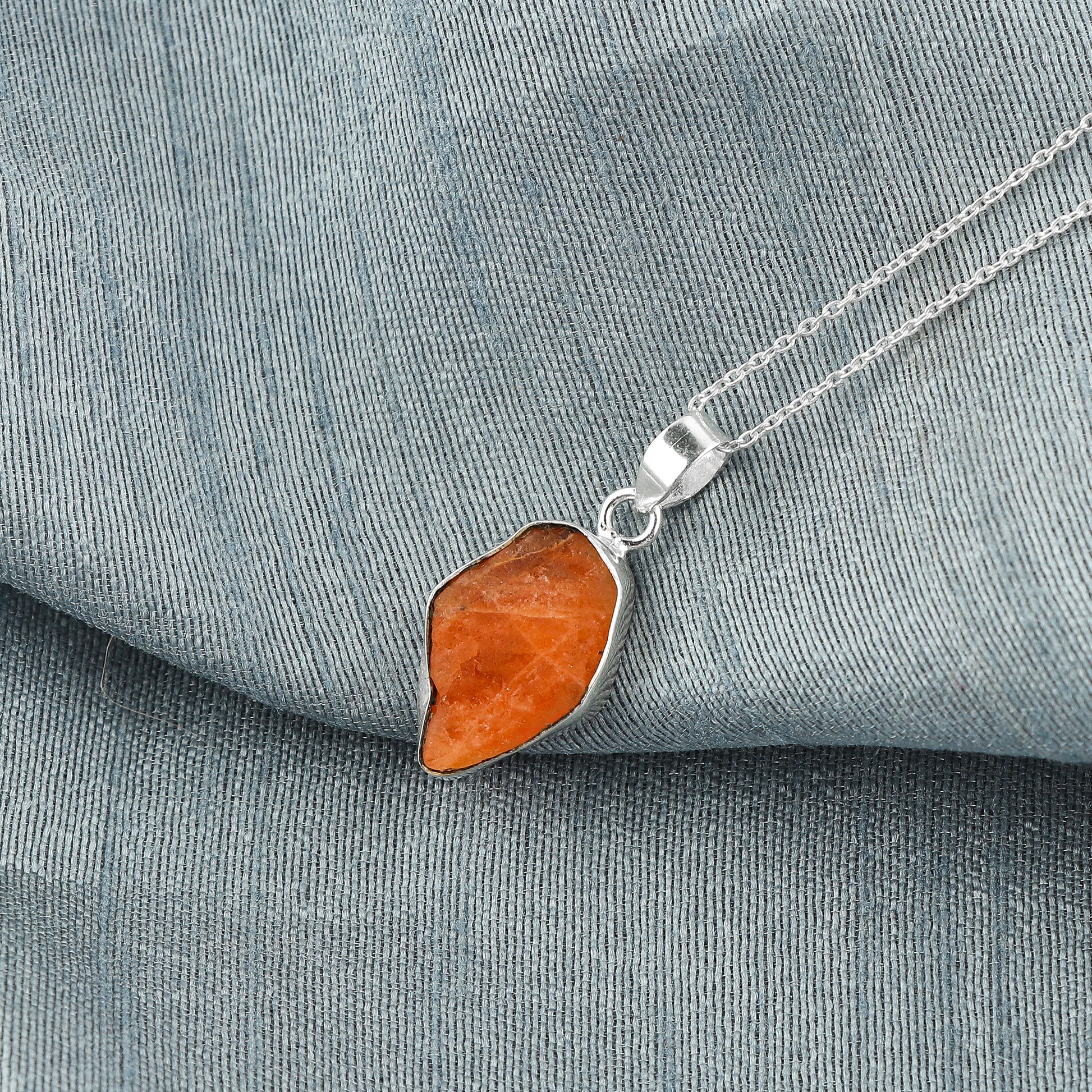 Triplite raw crystal pendant placed on grey fabric showing vibrant orange color, bezel setting and silver-plated chain details