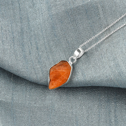 Triplite raw crystal pendant placed on grey fabric showing vibrant orange color, bezel setting and silver-plated chain details
