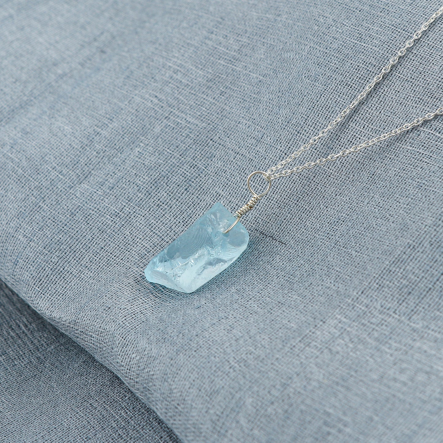 Sky blue topaz raw stone pendant placed on a soft fabric background highlighting its calming blue tone and silver bezel loop