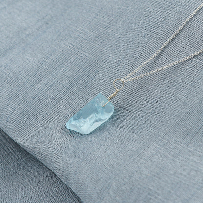 Sky blue topaz raw stone pendant placed on a soft fabric background highlighting its calming blue tone and silver bezel loop