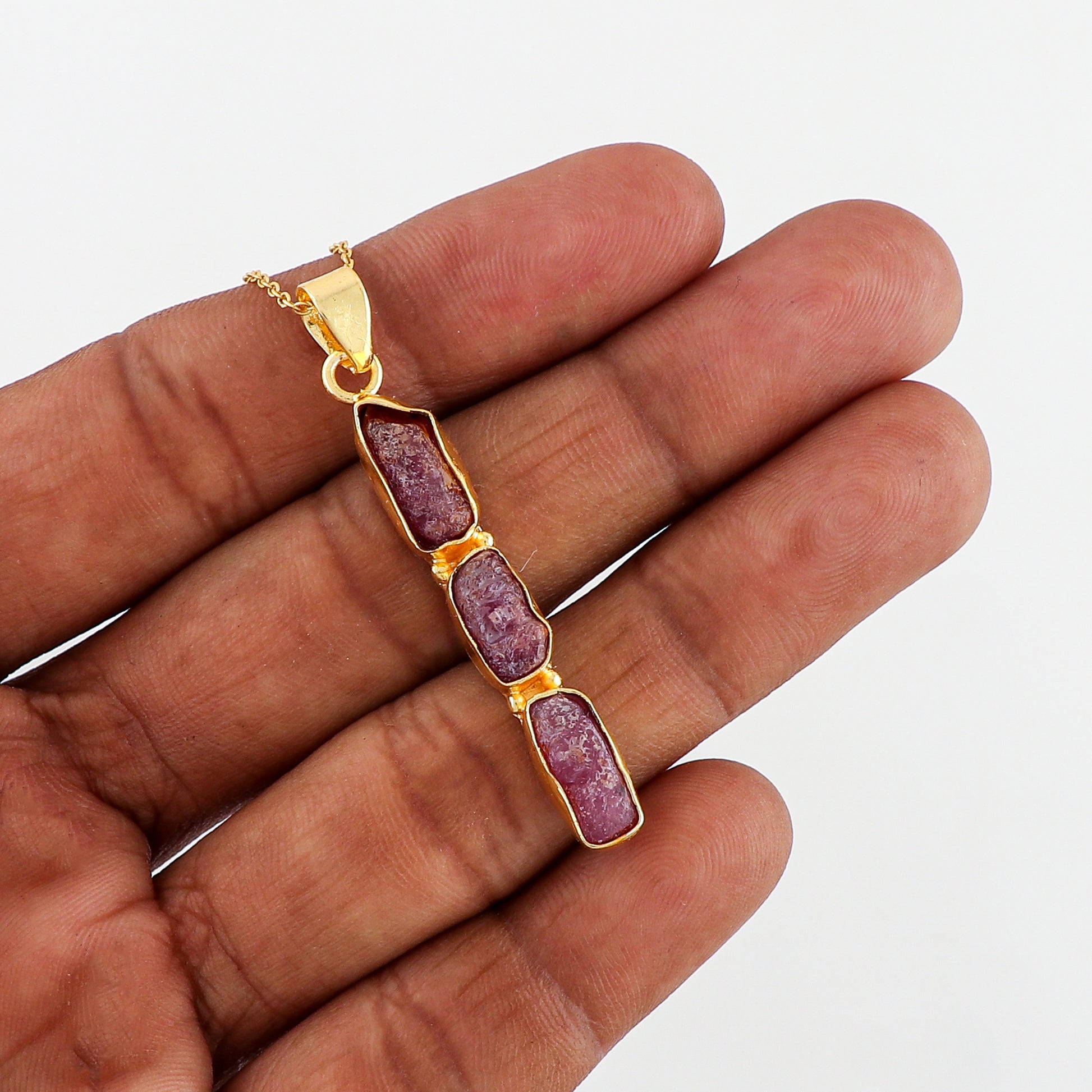 Natural pink ruby raw stone pendant held in hand showing irregular shape and gold-plated bezel on fine chain