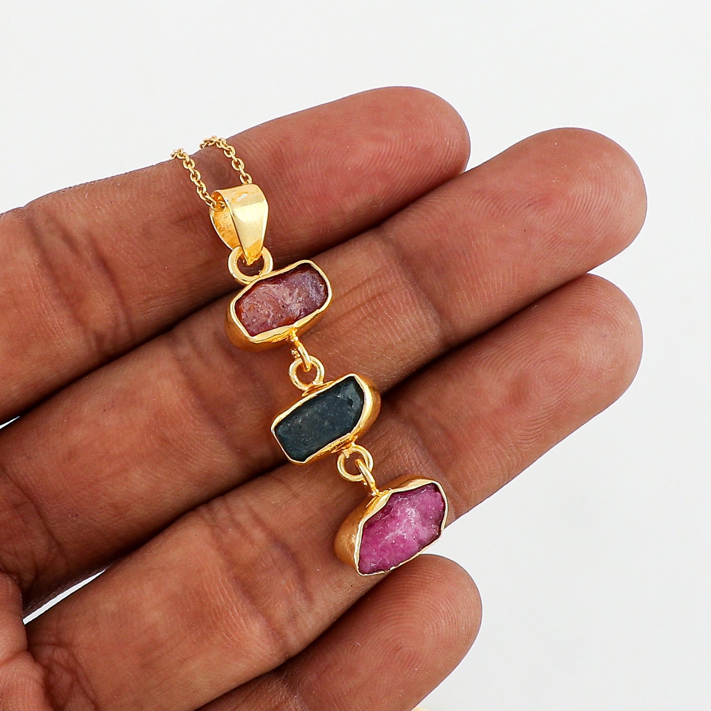 Natural sapphire and ruby raw stone pendant held in hand highlighting irregular shapes, dual gemstones and gold-plated bezel on fine chain